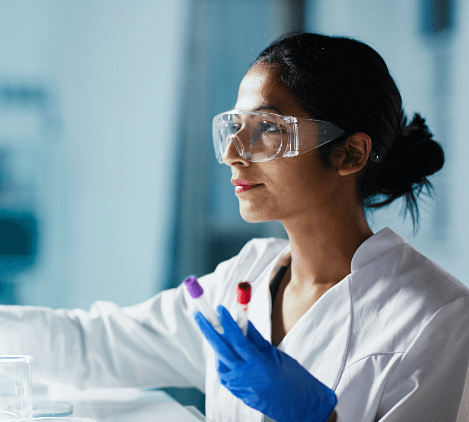 Woman in lab coat and safety goggles and gloves holds vials.